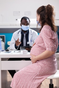 Lekarz przeprowadza konsultację lekarską z kobietą w ciąży, noszącą maseczki na twarz. kobieta spodziewająca się dziecka i otrzymująca poradę medyczną od lekarza przy biurku podczas pandemii covid 19