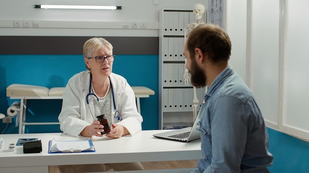 Lekarz rodzinny podający butelkę tabletek na wyleczenie chorego mężczyzny podczas wizyty kontrolnej w gabinecie lekarskim. Wyjaśnienie leczenia na receptę w kolbie z lekami podczas wizyty konsultacyjnej.