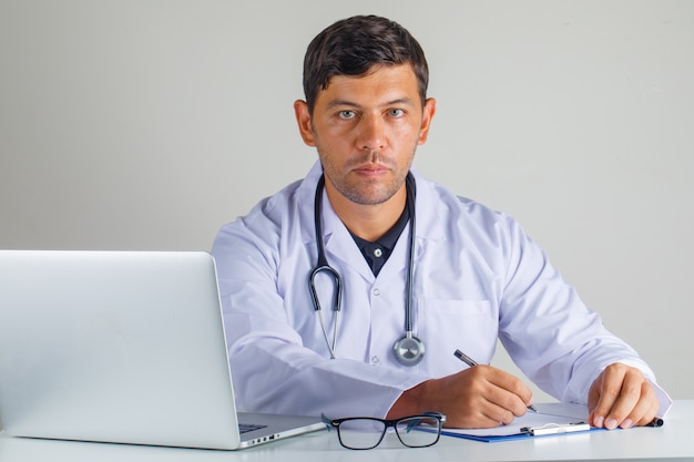 Lekarz siedzi i robienie notatek w białym fartuchu i stetoskopie