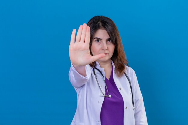 Lekarz w średnim wieku ubrany w biały fartuch i stetoskop stojący z otwartą ręką robi znak stopu z poważnym i pewnym siebie gestem obrony na niebieskim tle