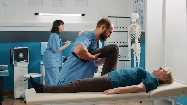 Lekarz wykonujący ćwiczenia podnoszące nogi w celu rozprostowania nóg starego pacjenta z zaburzeniami mechanicznymi w gabinecie rehabilitacyjnym. Specjalista osteopatii wykonujący zabieg fizykoterapii w celu wyleczenia kobiety.