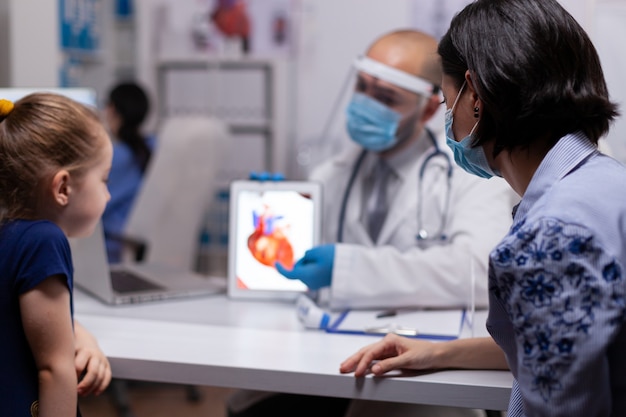 Lekarz z maską ochronną i wizjerem opowiada o pracy serca z pacjentami korzystającymi z tabletu. specjalista w leczeniu chorób noszący sprzęt ochronny podczas globalnej pandemii
