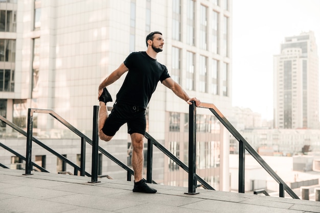 Lekkoatletycznego mężczyzna rozciągania nóg przed treningiem na świeżym powietrzu. biegacz w czarnej odzieży sportowej ćwiczenia o poranku trzymając poręcz. aktywne życie. rano miasto na tle. rano rozgrzewka.