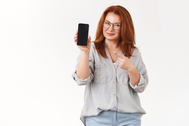 Lepiej sprawdź siebie Pewność sukcesu, rudowłosa kobieta w średnim wieku, dzieląca się tajemnicami zawodowymi, trzymaj smartfon, wskazując ekran telefonu, promuj aplikację, uśmiechając się, zachwycony Koncepcja technologii