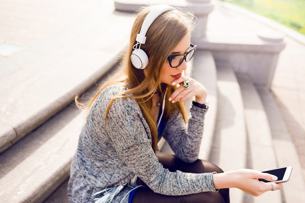 Letni styl życia słoneczny obraz ładnej młodej blondynki kobiety słuchającej muzyki przez słuchawki, trzymając telefon komórkowy, siedząc na ulicy, marzy. Noszenie stylowego wiosennego stroju.