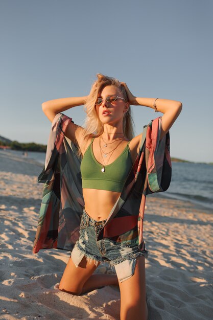 Letnie zdjęcie sexy blond kobieta w zielony krótki top i dżinsy, pozowanie na tropikalnej plaży.