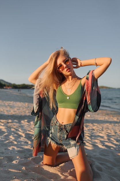 Letnie zdjęcie sexy blond kobieta w zielony krótki top i dżinsy, pozowanie na tropikalnej plaży.