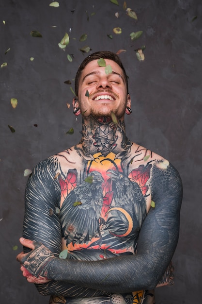 Bezpłatne zdjęcie liście spada nad uśmiechniętym tatuującym młodym człowiekiem z rękami krzyżować