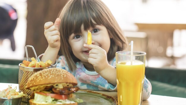 Little cute girl jedzenie kanapki fast food z frytkami i sokiem pomarańczowym w kawiarni. Koncepcja fast food.