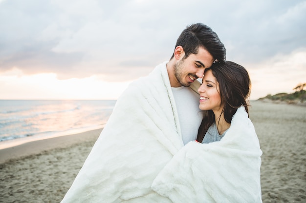 Bezpłatne zdjęcie loving para siedzi na plaży objętych białym kocem