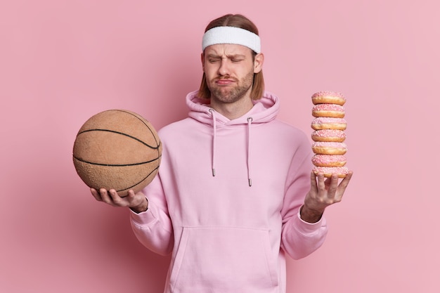 Ludzie sport koncepcja fast foodów. Niezadowolony wysportowany mężczyzna patrzy nieszczęśliwie na piłkę do koszykówki trzyma stos słodkich apetycznych pączków, nie chce się bawić ubrany w bluzę z kapturem.