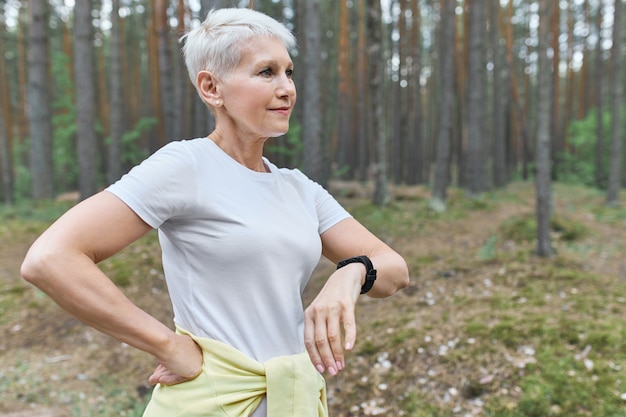 Ludzie, sport, zdrowie i technologia. Aktywna emerytowana kobieta nosząca inteligentny zegarek do śledzenia jej postępów podczas ćwiczeń cardio na świeżym powietrzu.