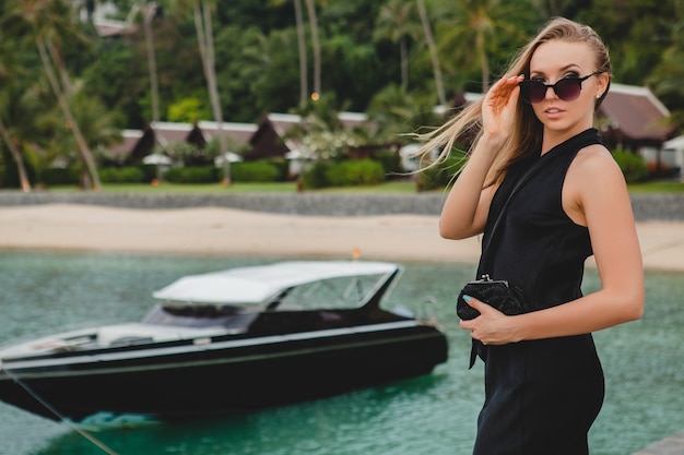 Bezpłatne zdjęcie luksusowa seksowna atrakcyjna kobieta ubrana w czarną sukienkę, pozowanie na molo w luksusowym hotelu, na sobie okulary przeciwsłoneczne, wakacje, tropikalna plaża
