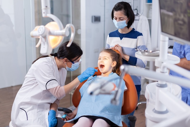 Lustro kątowe używane przez lekarza dentysty na dziewczynce z otwartymi ustami w gabinecie stomatologicznym. Lekarz stomatolog podczas konsultacji jamy ustnej dziecka w gabinecie stomatologicznym z wykorzystaniem nowoczesnych technologii.