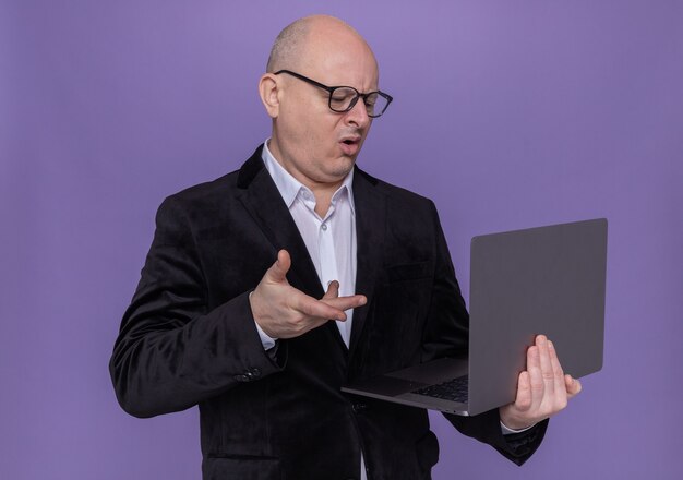 Łysy mężczyzna w średnim wieku w garniturze w okularach trzymając laptopa patrząc na ekran, jest zdezorientowany i niezadowolony, stojąc nad fioletową ścianą