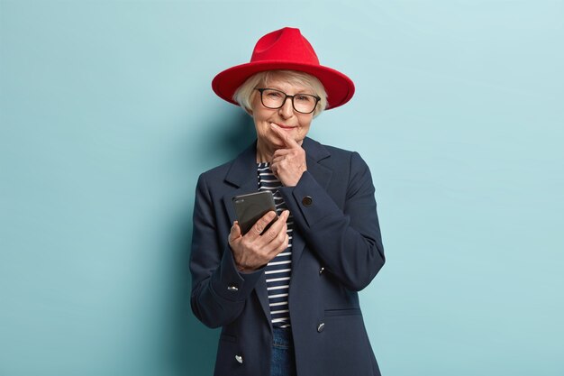 Mądra, przemyślana, zadowolona starsza, pomarszczona kobieta trzyma podbródek, czyta powiadomienie, jest podłączona do bezprzewodowego internetu, nosi czerwone nakrycie głowy i modny płaszcz, otrzymuje rabat na e-maile. Ludzie, wiek, mądrość