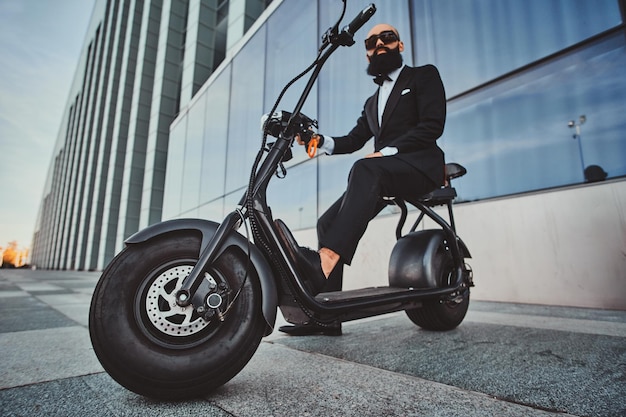 Mądry, atrakcyjny biznesmen siedzi na swoim skuterze elektrycznym, pozując dla fotografa.