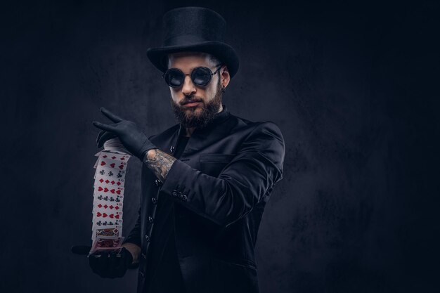 Bezpłatne zdjęcie magik w czarnym garniturze, okularach przeciwsłonecznych i cylindrze, pokazujący sztuczkę z kartami do gry na ciemnym tle.