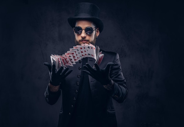 Magik w czarnym garniturze, okularach przeciwsłonecznych i cylindrze, pokazujący sztuczkę z kartami do gry na ciemnym tle.