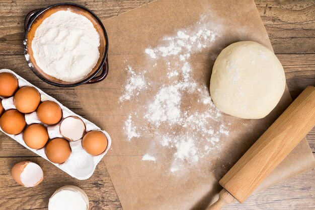 Mąka; karton jajek; ciasto; wałek do ciasta na pergaminie ponad drewnianym biurkiem