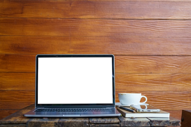 Makieta laptopa z pustym ekranem z notebookiem, filiżanką kawy i smartfonem na stole tła drewnianej ściany w kawiarni, biały ekran