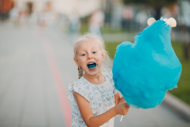 Mała blond dziewczynka jedzenie waty cukrowej i pokazuje niebieski język w parku.