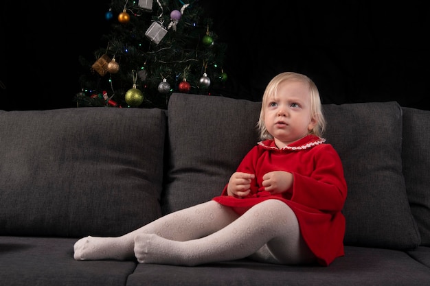 Mała blondynka w czerwonej sukience siedzi na kanapie na tle choinki i czarnej ściany. wigilia. nowy rok