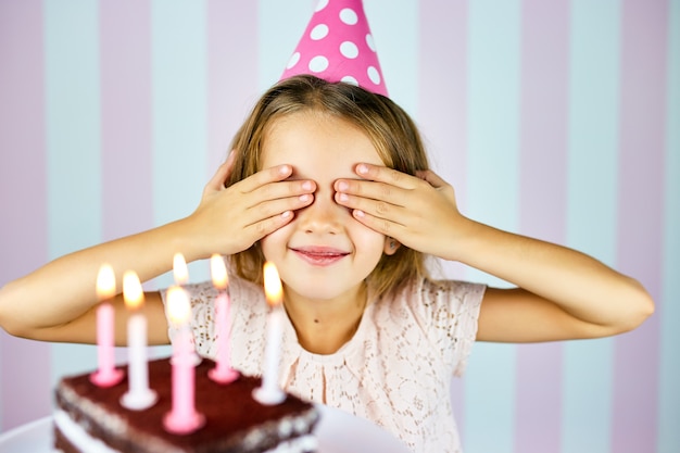 Mała blondynka w różowej czapce urodzinowej uśmiecha się, zamyka oczy, składa życzenie, zaskakuje czekoladowy tort ze świeczkami. dziecko obchodzi swoje urodziny.