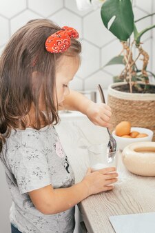 Mała ciemnowłosa dziewczynka 3 lata w czerwonej opasce piecze szarlotkę w kuchni. dziecko wkłada cukier do miarki. dzieci pomagają w pracach domowych. dziecko gotowanie żywności. pionowy