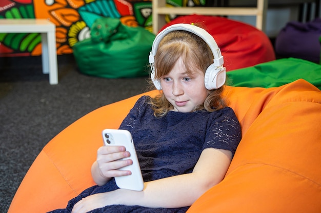Mała dziewczynka korzysta z telefonu i słucha muzyki przez słuchawki, siedząc na krześle z torbą
