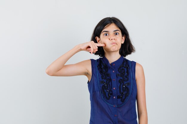 Mała dziewczynka naciskając palec na wydmuchiwanym policzku w niebieskiej bluzce i wygląda śmiesznie