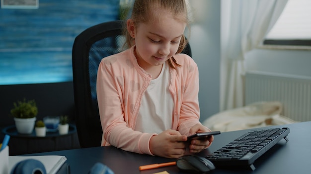 Bezpłatne zdjęcie mała dziewczynka patrząc na wyświetlacz smartfona i uśmiechając się do biurka. małe dziecko w szkole podstawowej za pomocą telefonu komórkowego z ekranem dotykowym do zdalnej lekcji online. kształcenie na odległość