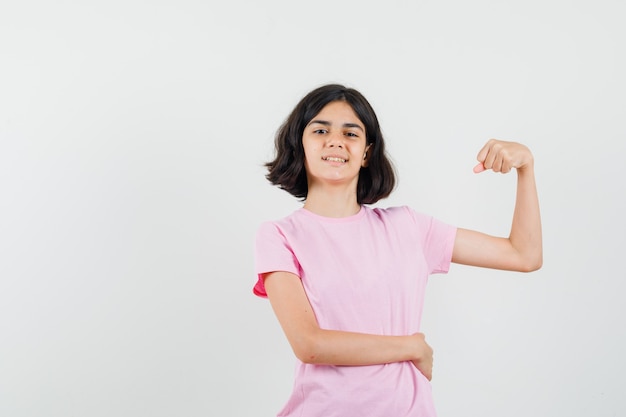 Mała dziewczynka pokazuje mięśnie ramion w różowej koszulce i wygląda pewnie, widok z przodu.