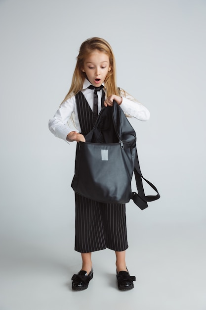 Mała dziewczynka pozuje w szkolnym mundurku z plecakiem na białej ścianie