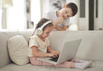 Mała dziewczynka pracuje z komputerem siedząc na kanapie, podczas gdy jej brat patrzy