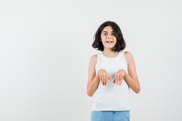 Mała dziewczynka robi zabawny gest w białą bluzkę, szorty i patrząc rozbawiony, widok z przodu.