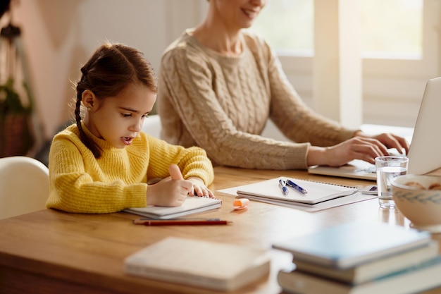 Mała dziewczynka rysująca na papierze, podczas gdy jej mama pracuje na laptopie w domu