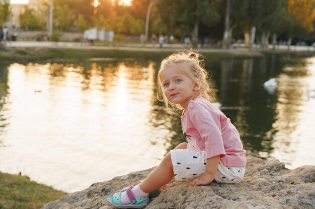 Mała dziewczynka siedzi na ogromnym kamieniu w parku w pobliżu jeziora