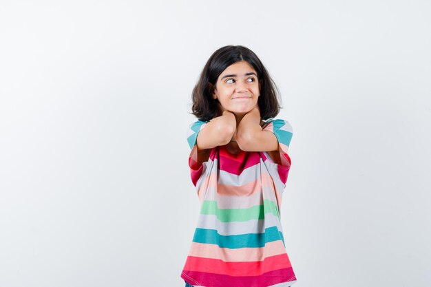 Mała dziewczynka trzymająca się za ręce na szyi, odwracająca wzrok w t-shirt, dżinsy i patrząca szczęśliwa, widok z przodu.