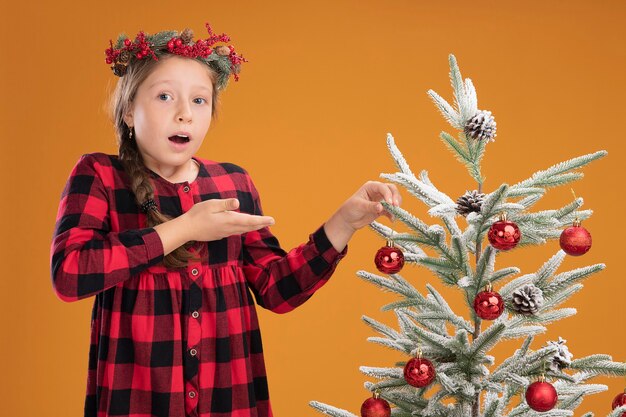 Mała Dziewczynka Ubrana W świąteczny Wieniec W Kraciastą Sukienkę Dekorującą Choinkę, Wyglądająca Na Zdezorientowaną I Zaskoczoną, Prezentując Rękę Stojącą Nad Pomarańczową ścianą