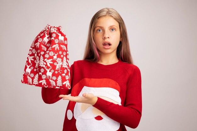 Mała dziewczynka w bożonarodzeniowym swetrze trzyma czerwoną torbę świętego mikołaja z prezentami, przedstawiając ją ramieniem w dłoni, patrząc zdezorientowaną, stojąc na białym tle