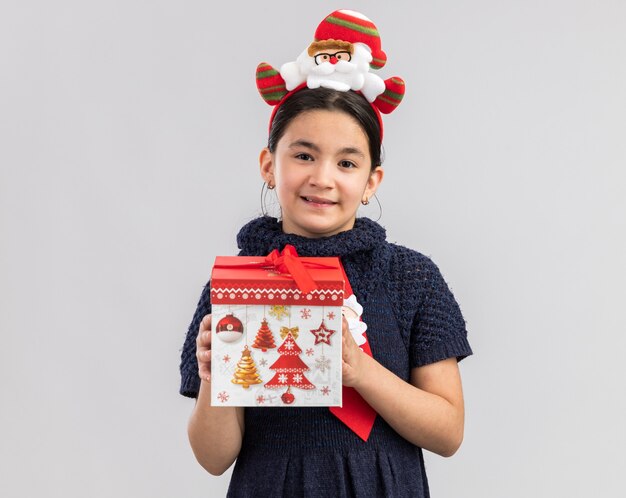 Mała dziewczynka w dzianinowej sukience na sobie czerwony krawat z zabawną świąteczną obwódką na głowie trzyma prezent na Boże Narodzenie patrząc z uśmiechem na twarzy szczęśliwy i pozytywny