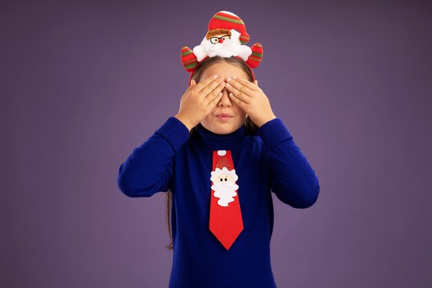 Mała Dziewczynka W Niebieskim Golfie Z Czerwonym Krawatem I Zabawną świąteczną Obwódką Na Głowie Zakrywającą Oczy Rękami Stojącymi Na Fioletowym Tle