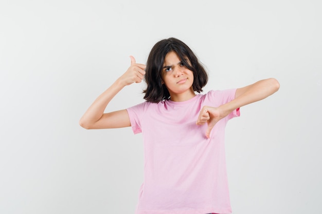 Mała dziewczynka w różowej koszulce pokazuje kciuki w górę iw dół i wygląda niezdecydowanie, widok z przodu.