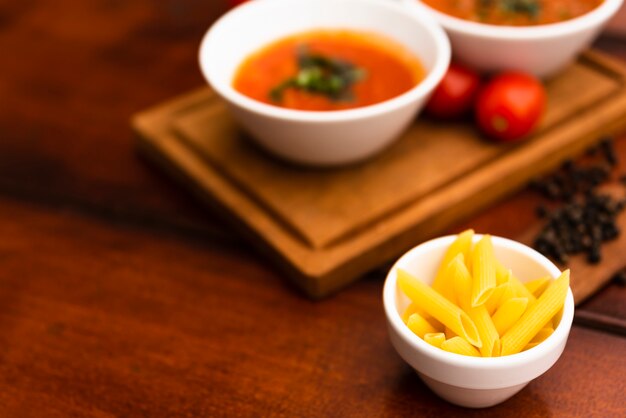 Mała miska surowego makaronu penne na stole z rozmytymi sosami i pomidorami na desce