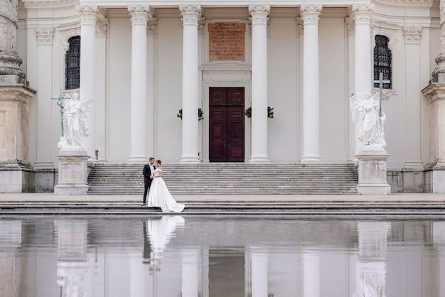 Mała para ślubna idzie blisko ogromnej katedry z białymi kolumnami i odbiciem w wodzie