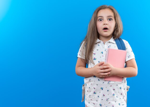 Mała piękna dziewczynka z długimi włosami z plecakiem trzymająca notatnik patrząca z przodu zdumiona i zdziwiona stojąca nad niebieską ścianą