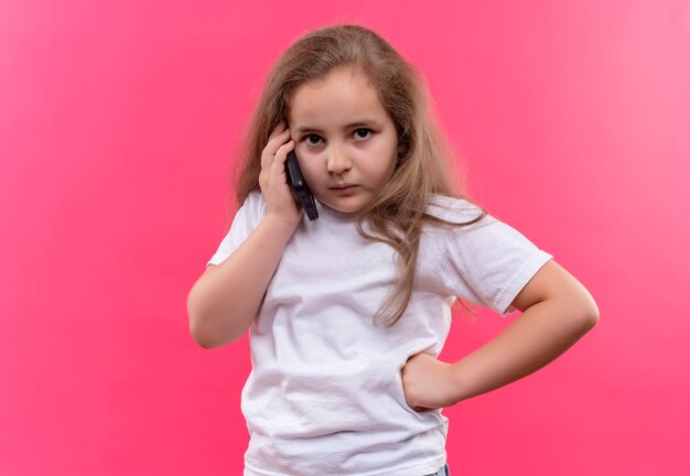 mała uczennica ubrana w białą koszulkę rozmawia przez telefon na na białym tle różowej ścianie