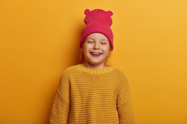 Mała urocza dziewczynka śmieje się radośnie, cieszy się z otrzymania nowej lalki od rodziców, zamyka oczy, bawi się w domu, nosi kapelusz z uszami i luźny sweter z dzianiny, odizolowany na żółtej ścianie.