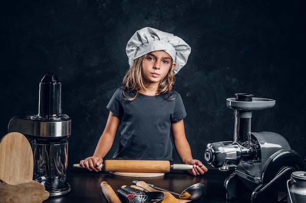 Mała Zamyślona Dziewczynka W Kapeluszu Szefa Kuchni Pozuje Fotografowi Z Wyposażeniem Kuchennym.